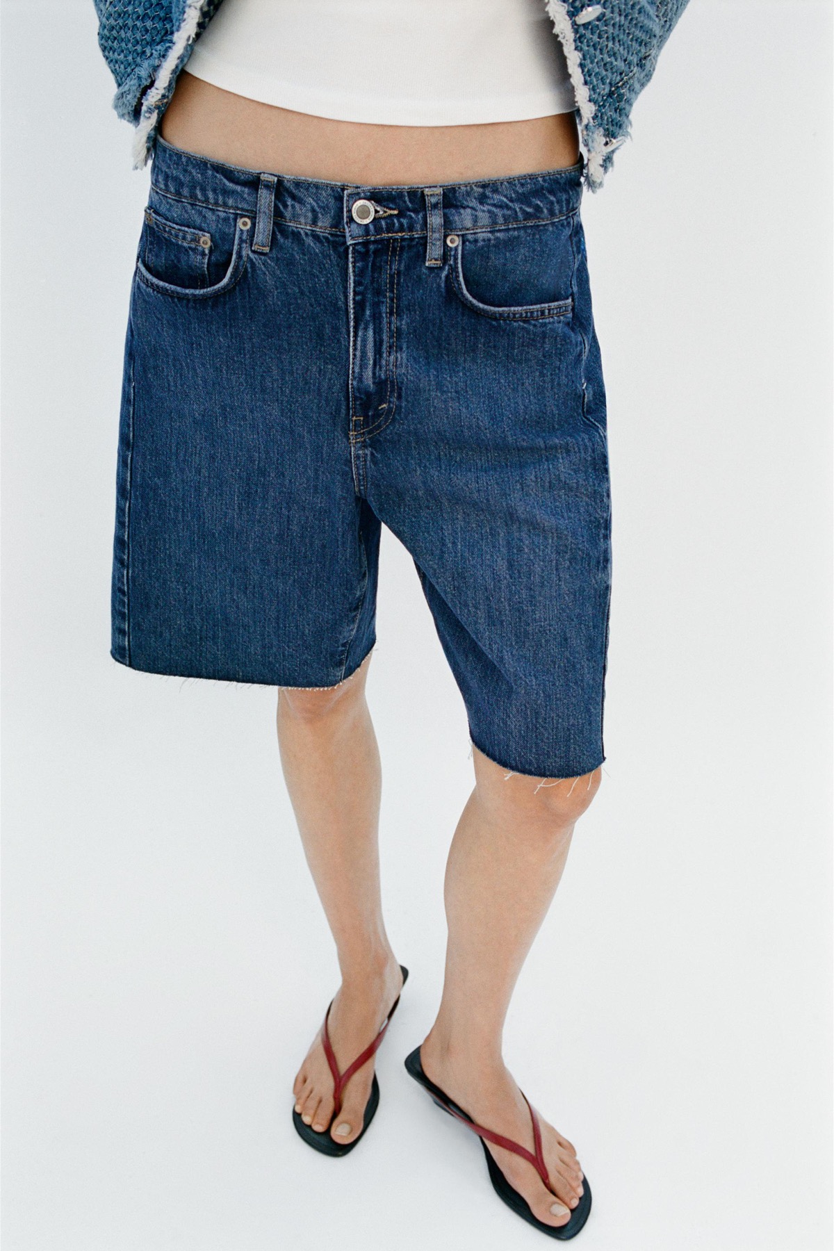 Close crop of dark-wash denim bermuda shorts, on-the-knee length, worn with flip-flops