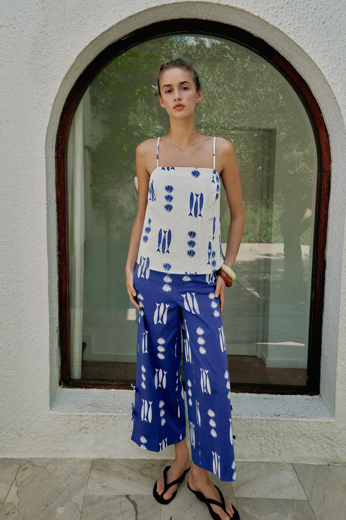 Woman wearing a white cami top and wide-leg trousers printed with a hand-drawn blue sardine and shell pattern