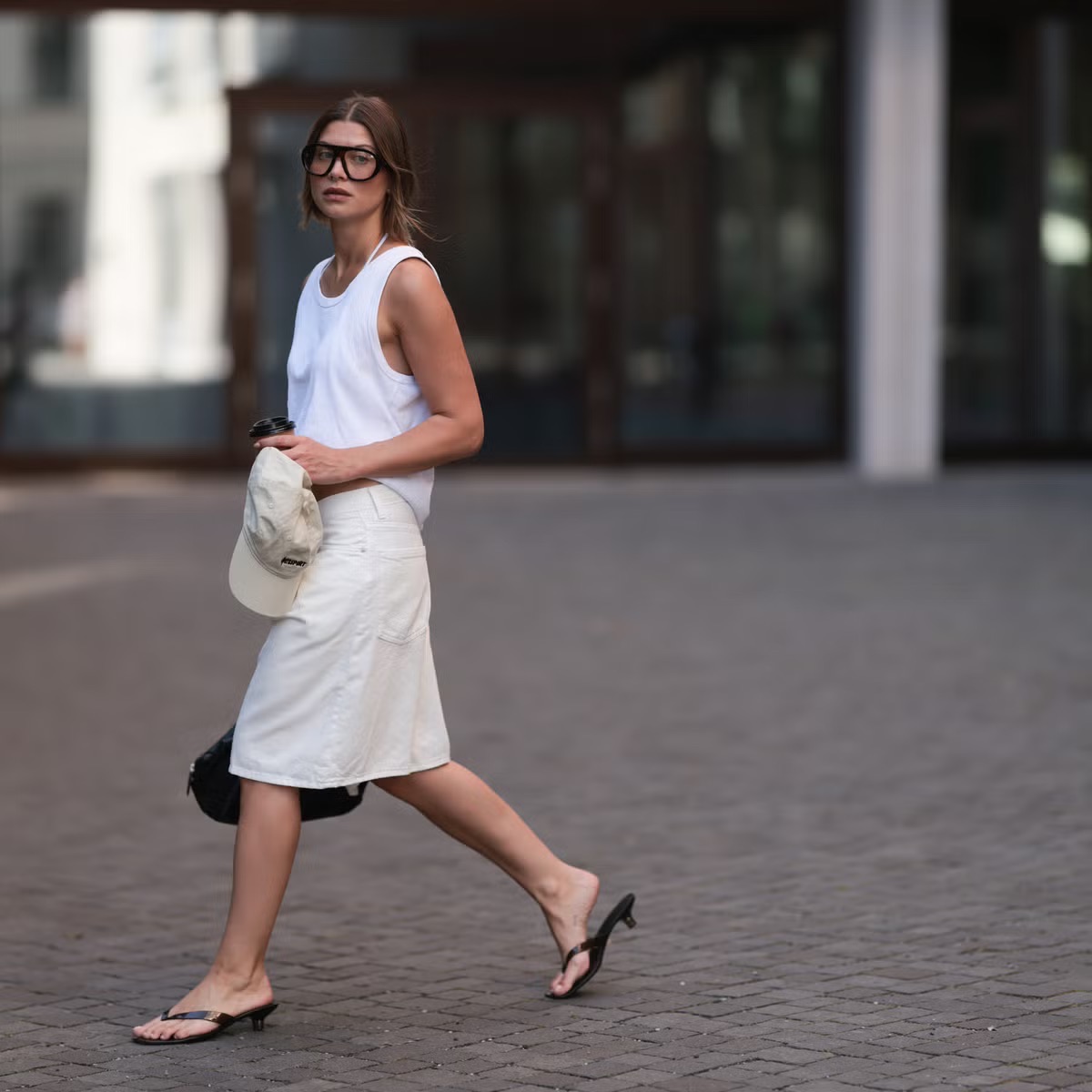 Street style shot of a woman in a white tank and white denim skirt, walking in black kitten-heel flip flops