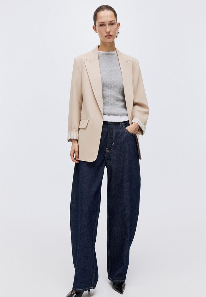 Woman wearing an oversized beige blazer over a grey knit and dark wide-leg denim, front view.