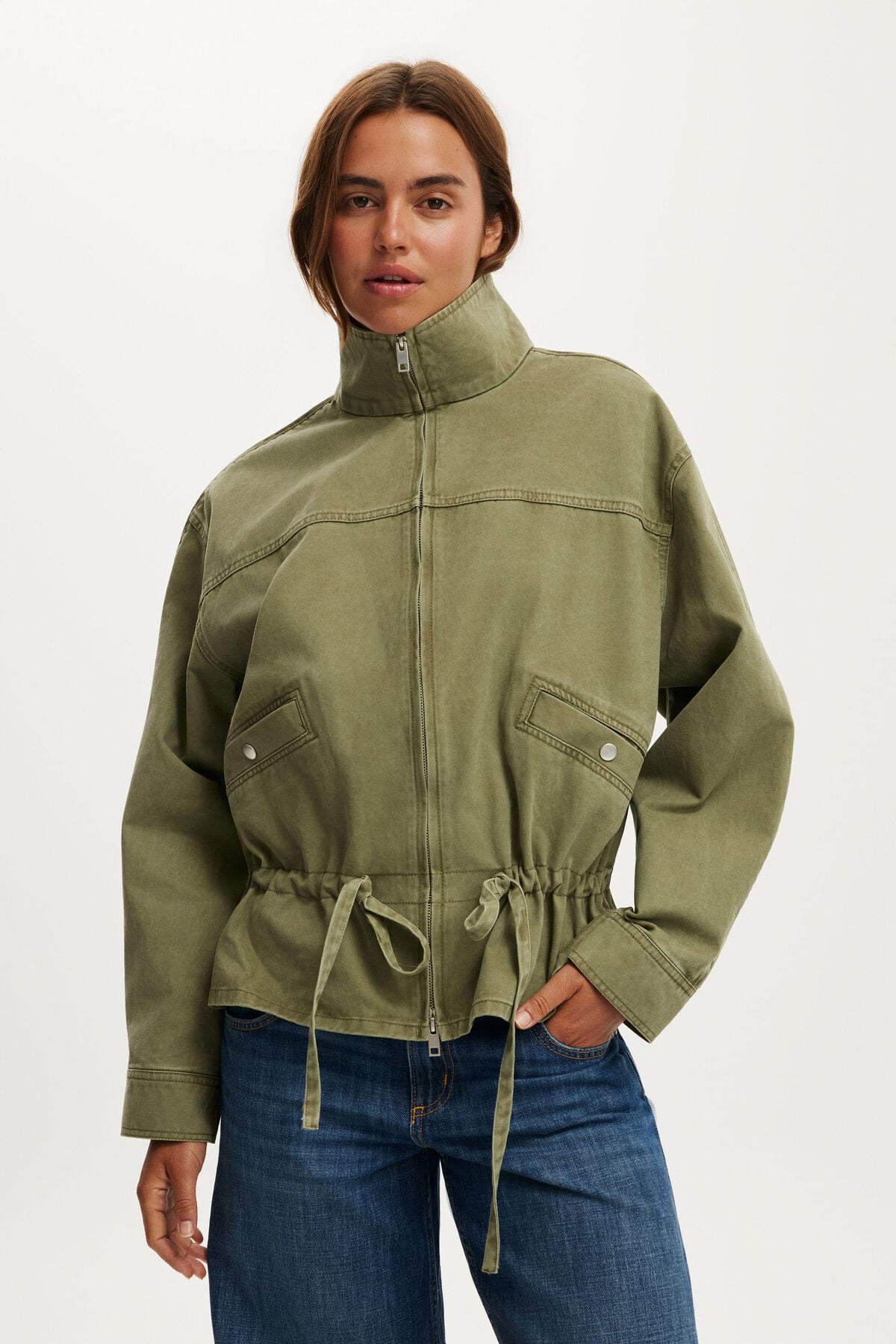 Woman wearing a khaki green funnel neck jacket with a drawstring waist, over blue jeans