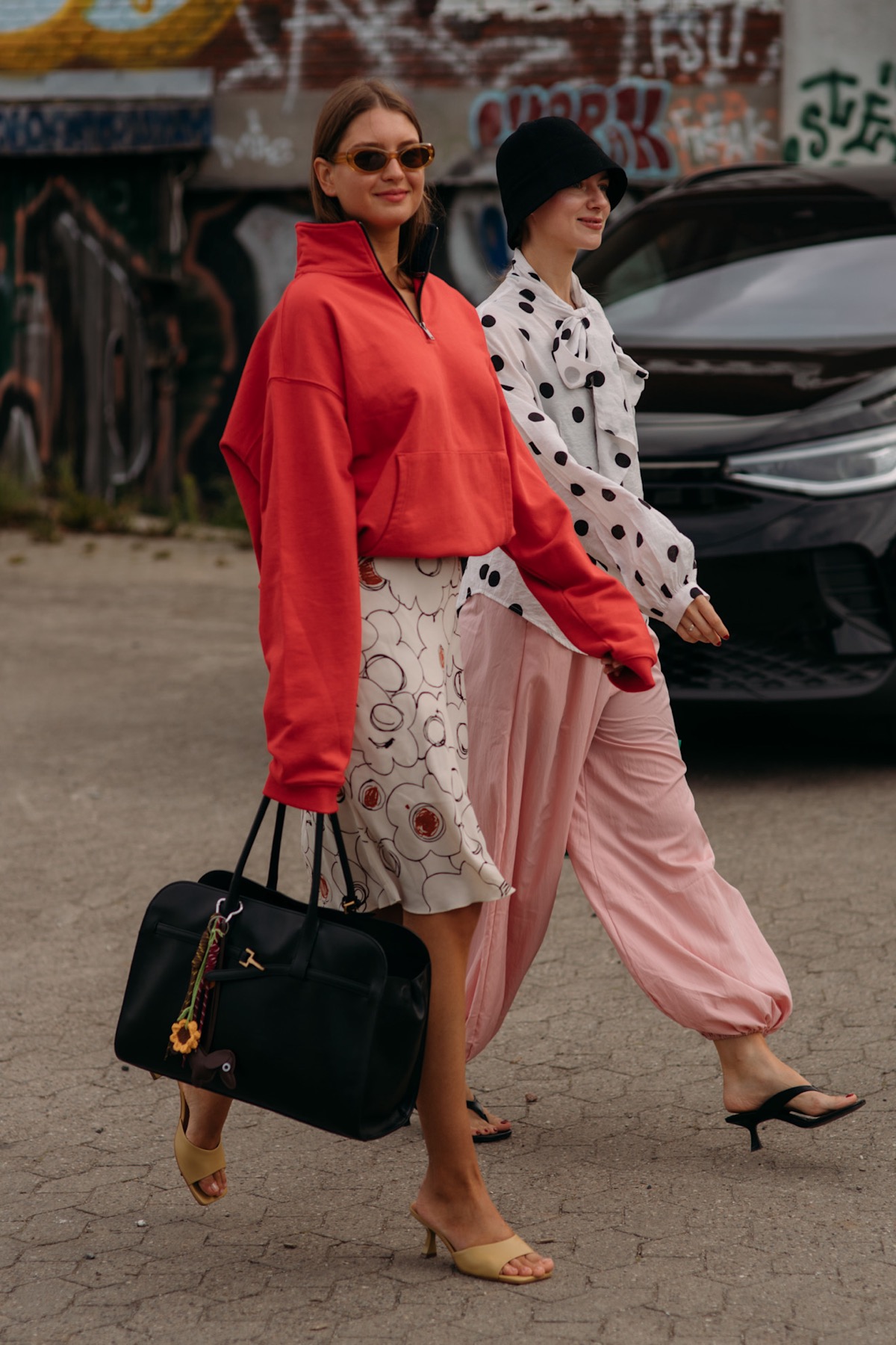 Street-style shot of two women, one in a red funnel-neck track jacket over a printed skirt, the other in a polka dot blouse and pink balloon pants