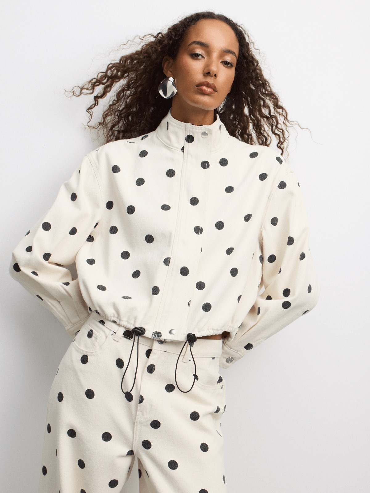 Woman wearing a cropped ecru denim jacket with black polka dots, funnel neck and drawstring waist